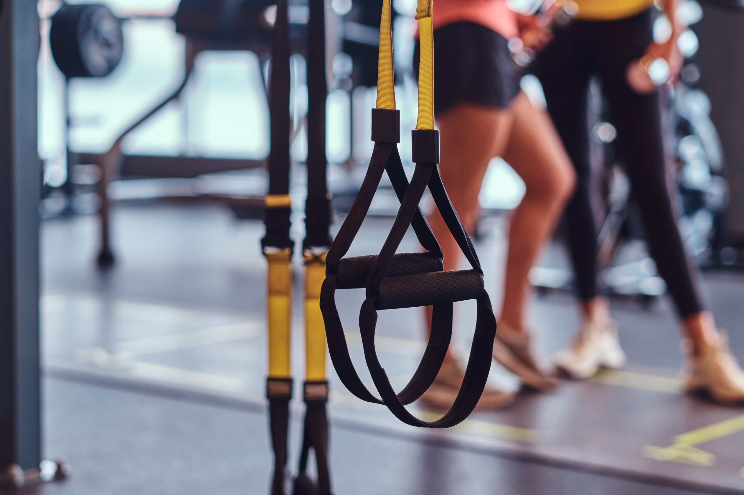 Sports suspension straps in the foreground and two fitness girls talking in the background in the modern gym.